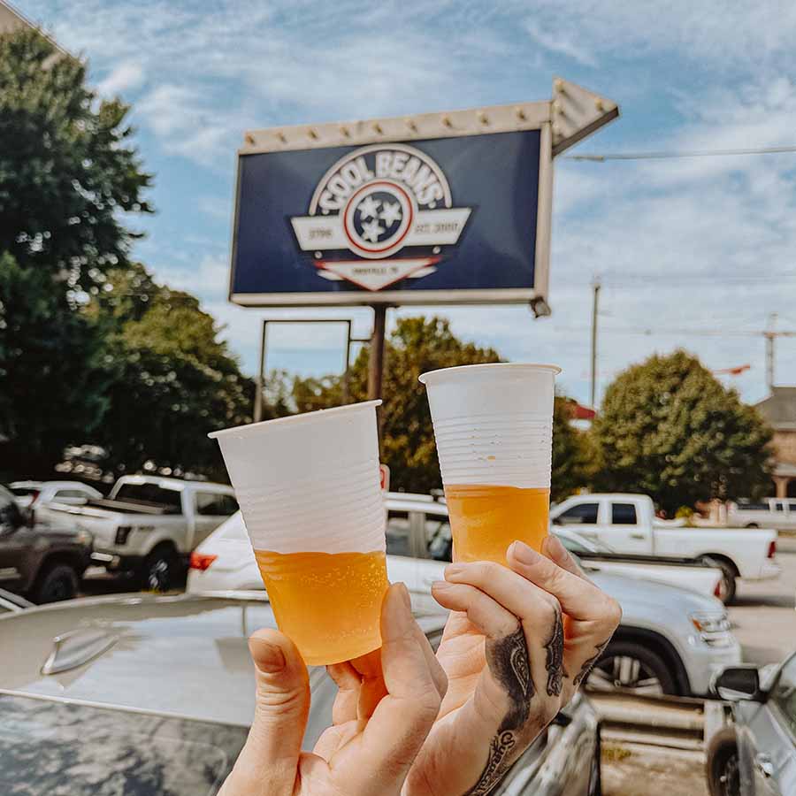 Two hands hold beers outside a Knoxville Tennessee Bar & Grill, Tool Bench sign in the background.